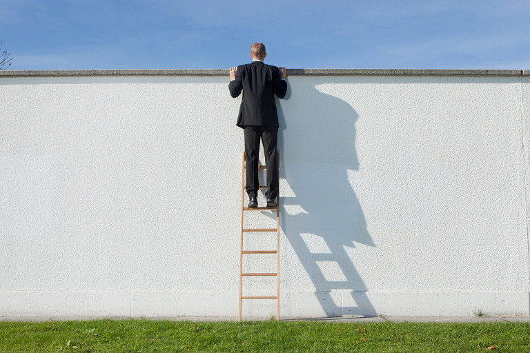 A man in a black suit climbs a wooden ladder to look over a tall white wall on a sunny day. His shadow is cast sharply on the wall, and green grass covers the ground below.