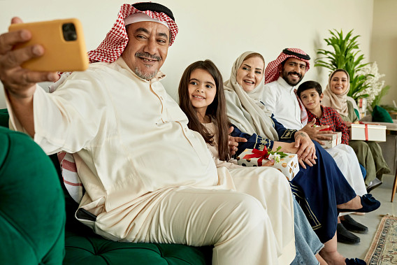 A family dressed in traditional attire is sitting together on a green couch, smiling as the elder man takes a selfie with a yellow phone. The group includes adults and children holding wrapped gifts, suggesting a festive or celebratory occasion. A bright and cheerful atmosphere is complemented by a plant in the background.