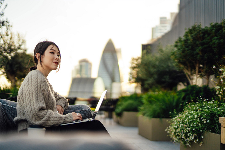 Young woman working on laptop outdoors, sitting on a bench in rooftop garden in London, surrounded by green plants.