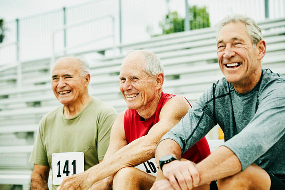Three senior men sitting on bleachers, smiling and enjoying themselves. Two are wearing numbered bibs, suggesting participation in a sporting event, while one is dressed in casual athletic wear. The scene is outdoors with a bright and cheerful atmosphere, emphasizing active and healthy aging.