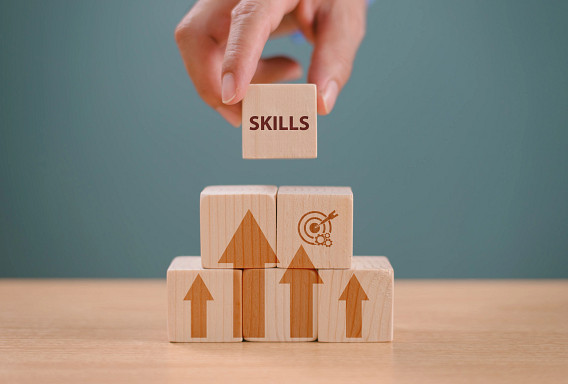 A hand places a wooden block labeled “SKILLS” on top of stacked wooden cubes with upward arrows and a target icon. The blocks sit on a table against a plain background, symbolizing growth, development, and goal-oriented learning.