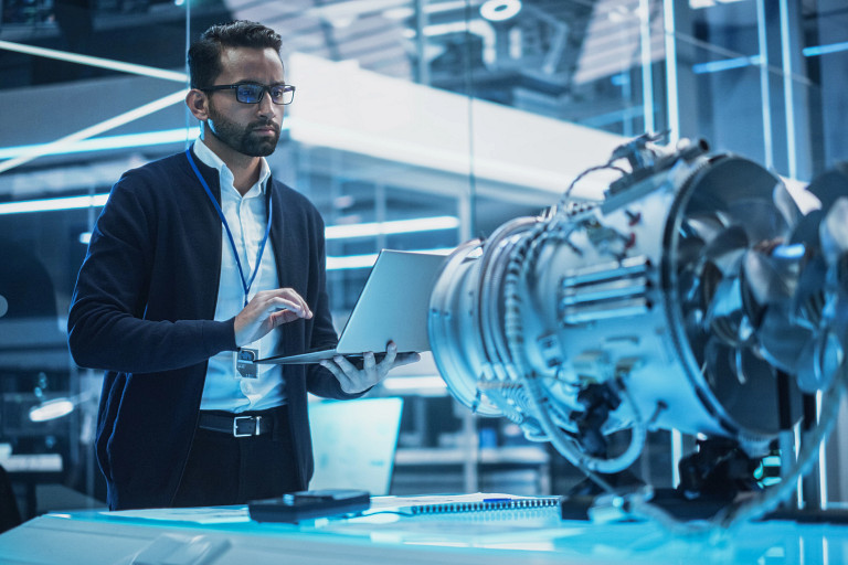 An Indian man in a modern, high-tech lab works on a laptop near a detailed turbine or jet engine model. The environment is illuminated with blue lighting, featuring glass walls, futuristic equipment, and a focused atmosphere.