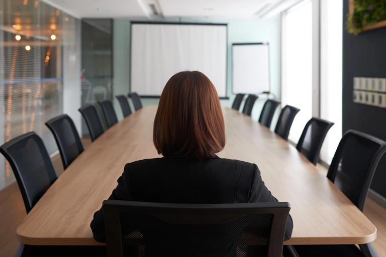 A person sits alone at the head of a long conference table, viewed from behind, in a modern meeting room. Empty chairs line both sides of the table, with presentation screens and large windows creating a quiet, formal atmosphere.