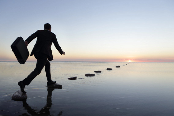 A businessperson carrying a briefcase steps across stones in shallow water toward the horizon at sunrise. The calm sea reflects the sky, and the line of stones leads forward, suggesting movement, progress, and balance.