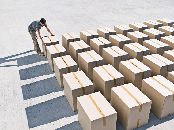 A person arranges sealed cardboard boxes laid out in neat rows on a large concrete surface, viewed from above. The boxes are evenly spaced, creating a grid pattern with long shadows cast in bright sunlight.