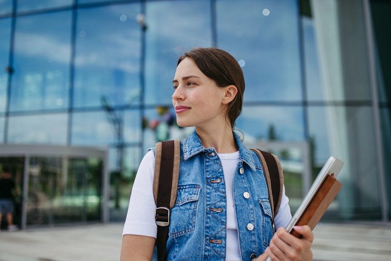 Portrait of young, pretty female student in front of university building or campus, backpack on back. Holding textbook